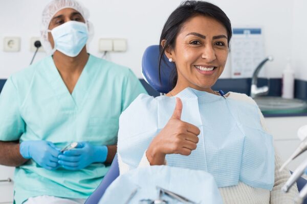 Happy latin american woman sitting in dental chair after teeth cure in modern clinic, gesturing thumbs up